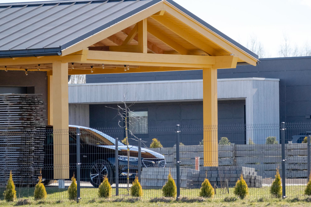 A yellow wooden carport with a car parked under it and small fir trees in the foreground